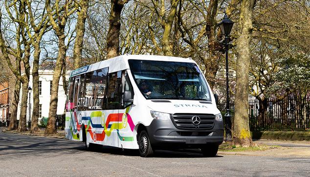 The image shows a white minibus with a Mercedes-Benz logo on the front, driving along a tree-lined street. The vehicle is marked with vibrant, colourful stripes in hues of green, yellow, orange, pink, and blue along its side, adding a lively contrast to the otherwise neutral colour of the bus. Large windows dominate its side, offering a clear view of the interior. The word "STRATA" is visibly printed on the side of the bus. In the background, bare tree branches suggest a setting either in late autumn or early spring, casting intricate shadows on the pavement. A row of buildings with classic architecture is partially visible through the trees.