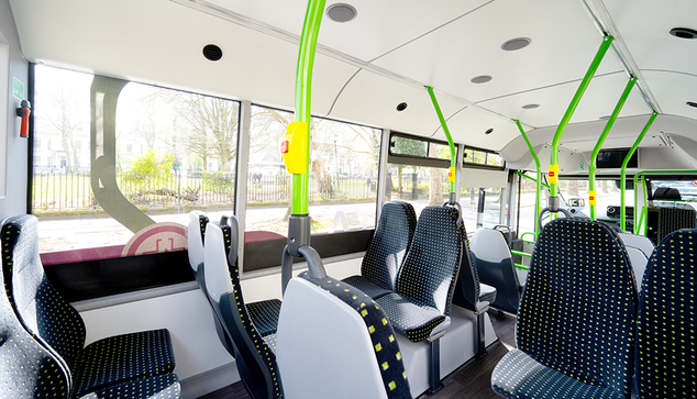 The image depicts the interior of a modern bus, focusing on the seating arrangement. The seats are upholstered in black fabric with white and yellow polka dot patterns. Several rows of seats are visible, with some facing forwards and others placed opposite each other. The bus features large windows allowing natural light to fill the interior. Overhead, a clean, white ceiling includes small circular lights and ventilation units. Bright green and yellow poles provide support for standing passengers and feature buttons for stop requests. The view outside the windows reveals a blurred landscape with trees and a fence, suggesting the bus is in motion or parked near a park.