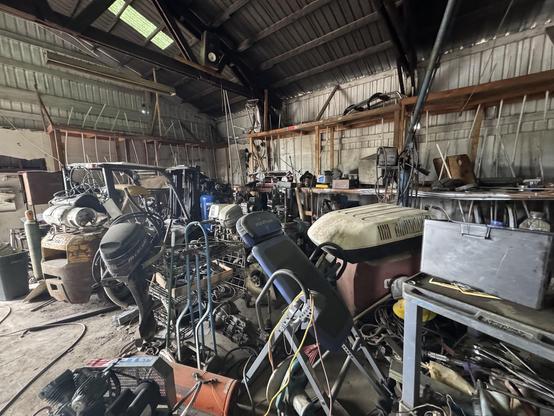 An cluttered workshop filled with various tools, machinery, and equipment. Items include engines, metal parts, and work benches, all organized in a disorganized manner under a high ceiling. Natural light comes in through skylights.