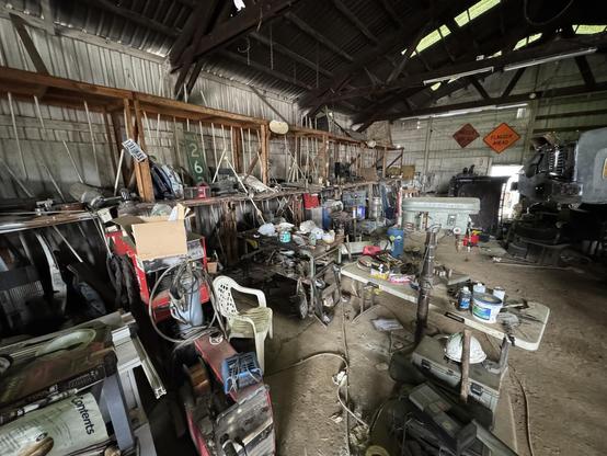A cluttered workshop interior featuring various tools and equipment, including workbenches, machinery, and scattered items. The space has a rustic wooden structure with visible metal shelves and overhead beams. Several warning signs are visible on the walls.