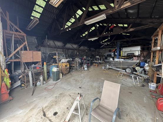 An industrial workshop or garage featuring various tools, equipment, and a boat. The space is cluttered with workbenches, machinery, and an overhead vehicle lift. Bright green light filters through skylights in the roof. A chair is visible in the