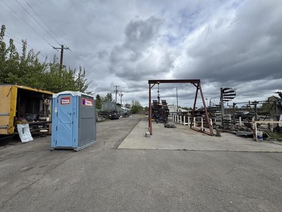 An industrial area featuring a portable toilet, a yellow truck, and various machinery and equipment along a concrete path. The sky is overcast with clouds, and there's greenery visible on the left side.