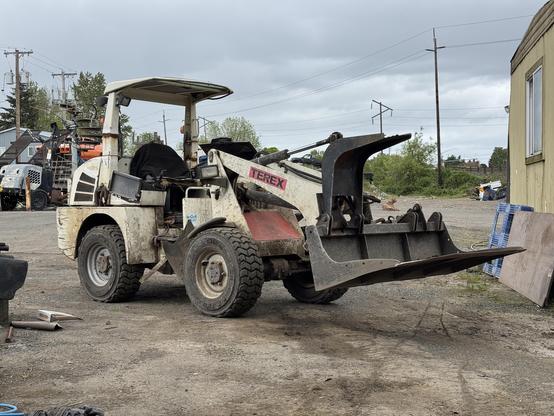 A dirty white Terex skid steer loader is parked on a gravel surface. The loader has a raised bucket at the front and is surrounded by various construction debris and equipment. In the background, there are power lines and additional machinery.