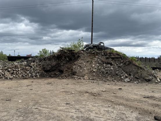 A pile of rubble with an abandoned, rusted vehicle perched on top. The sky is overcast with gray clouds, and there are some trees and power lines in the background.
