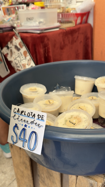 Fotografía a color de una bandeja de color azul con hielo y sobre del hielo pequeños botes con tapa que tienen una pegatina sobre la tapa que dice gracias. Un letrero al frente que dice con letras azules CARLOTA DE limón $40.00