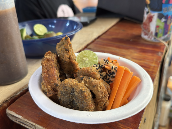 Fotografía a color de un plato con NOT-Calamar hecho con berenjenas capeadas acompañadas de not-mayo, ajonjolí negro, zanahorias en julianas y un limón partido a la mitad. Al derredor de la mesa hay un cajete con limones y una salsa en un bote.