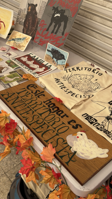 Fotografía a color de una mesa donde hay varios productos en venta. Una tabla de madera que dice: Este hogar es TERRITORIO ANTIESPECISTA. Unas camisetas color beige con la frase territorio antiespecista y una gallina dibujada en el medio. Otra playera color beige con la frase en letras negras. Al costado izquierdo unos afiches y dibujos de aves, pegatinas.