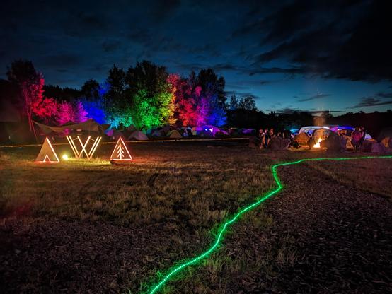 Menschen sitzen am Lagerfeuer und genießen das VVoid Camp. Im Hintergrund werden Bäume beleuchtet.