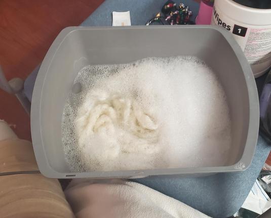 Gray basin with water, suds and white lace scarf in it, sitting on a chair with clutter