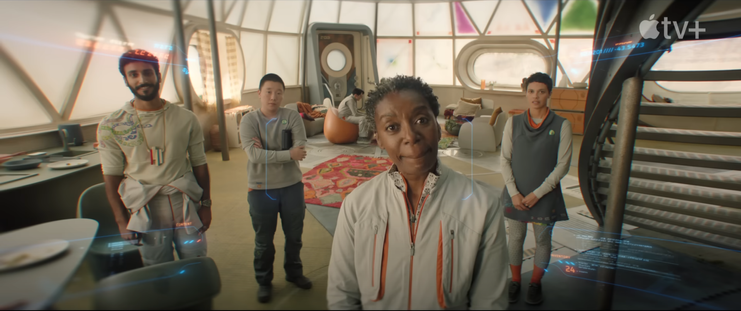 Murderbot's POV camera view showing mensah, pin-lee, and two others looking at murderbot in a building with translucent exterior coverings with some triangular panels painted either purple,blue or green. Mensah is looking quizzically. one of the others is looking happy. their outfits have light grey and orange accents