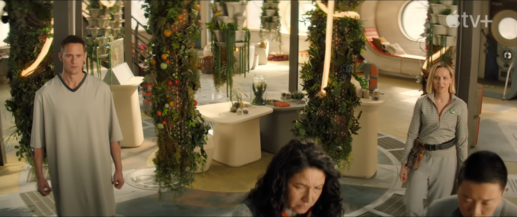 over the top shot of a room with greenery(fruits/vegs etc) attached to pylons hanging from the ceiling
a curve narrow staircase is in the backgroun, murderbot is not in its armour wearing a hospital gown on the left, two characters are cut off only seeing their head in the bottom right (one of them pin-lee), they're both working on something, a character with blonde hair and a tool belt with a green badge of their shirt is about to speak. much of the interior design is earthy by sparse in detail and not industrial.