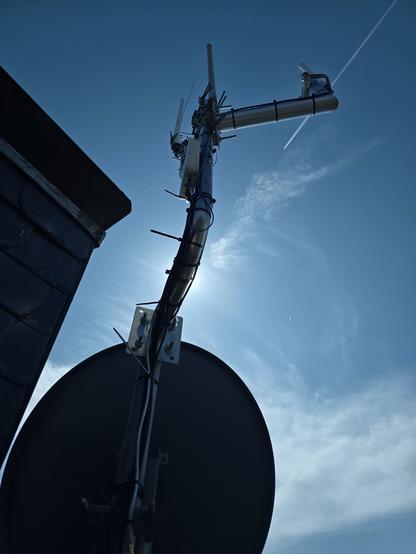 Gegen blauen Himmel im Vordergrund ein Antennenmast mit SAT Schüssel und zahlreichen Antennen (Adsb, AIS, Wettersonden, Lorawan), die an Ausleger-Masten hängen. Neue Antennenkabel mit Kabelbindern, die noch nicht abgeschnitten sind.