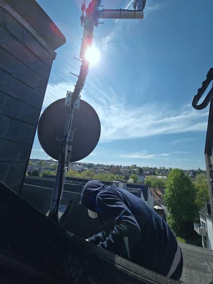 Foto aus einer Dachluke blauer Himmel grün er Horizont, im Vordergrund eine Person im Hoodie, die in der Dachfläche Kabel sortiert