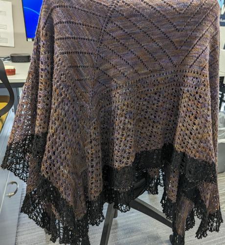 A lace shawl hanging on a chair. It looks largely brown, but with variegation. The section at the top has lines of lace "holes". The bottom section is much more lacy. There is an inch or two of black around the bottom edge.