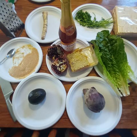 A Passover seder with parsley, horseradish root, charoset, an avocado, a beetroot, romaine lettuce, matzo on the side, and a center plate with a pomegranate, honey comb, and peach flavored sparkling grape juice