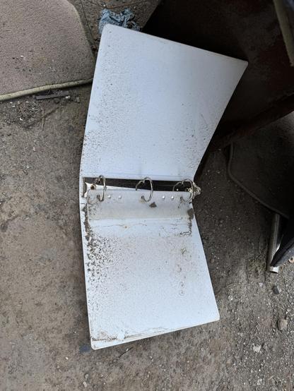 An open, dirty white binder lying on a gravel surface. The binder has metal rings for holding pages, but is empty and shows signs of wear.