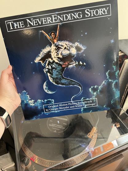 My hand holding the vinyl record for the Neverending Story soundtrack while the album plays below on a turntable. The cover image is Atreyu, the boy warrior riding Falkor the luck dragon, with the night sky and moon behind them