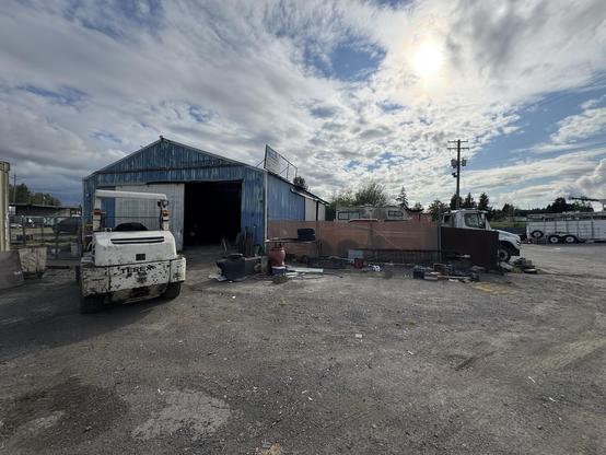 A gravel lot features a blue metal building, with a Terex vehicle parked in front. There are various items scattered around, including containers and tools, under a partly cloudy sky. A white truck is visible in the background along with a power pole.