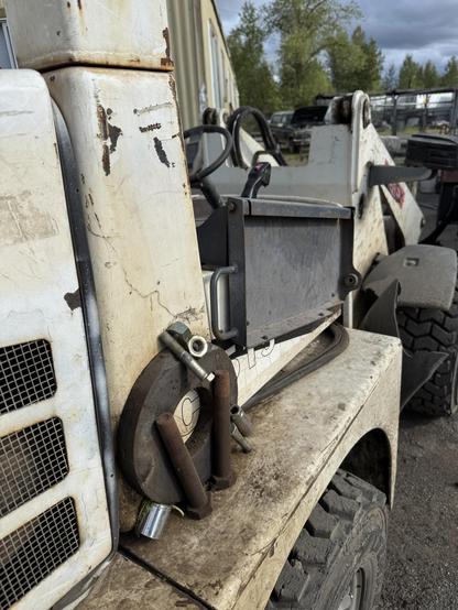 A close-up of a dirty, white machinery component, likely part of a skid steer loader. It shows rusted tools and machinery parts, with a blurred background of other equipment and trees.