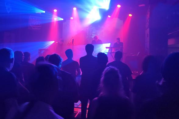 The industrial band Front Line Assembly performing on stage, surrounded by pink and blue stage lights. A silhouetted crowd stands in the foreground: people watching the band's performance.