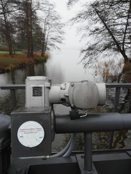 The image shows a mechanical device or machinery part near a body of water, possibly related to water management or a facility. The background includes trees and a serene, misty river or lake scene.