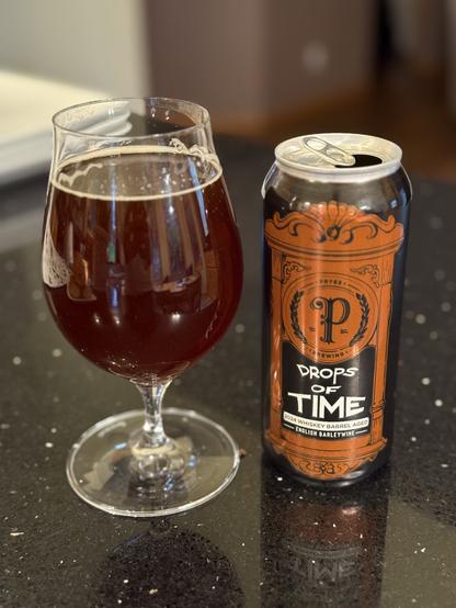 A snifter of ruby red beer next to a black can with a shiny orange front with lettering in white and black. The Pryes scripted “P” logo is front and center