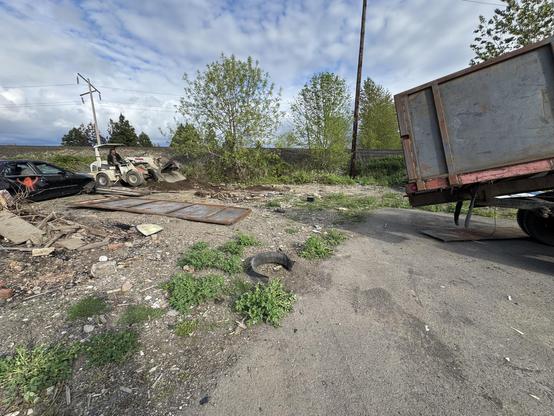 A construction site with debris and overgrown vegetation. A small machinery is operating near a black car and an overturned truck. Power lines are visible in the background under a cloudy sky.