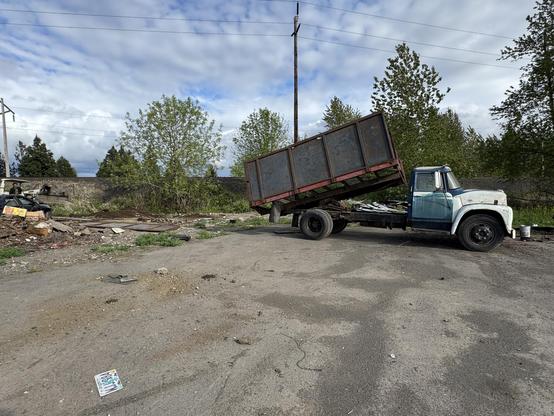 An old truck with a raised dump bed is parked on a dirt lot, surrounded by debris and overgrown vegetation. Power lines and a cloudy sky are visible in the background.