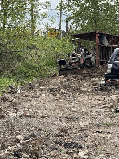 A construction site features a person operating a small excavator on a gravel and debris-strewn path. In the background, trees surround the area, with a train visible on elevated tracks.