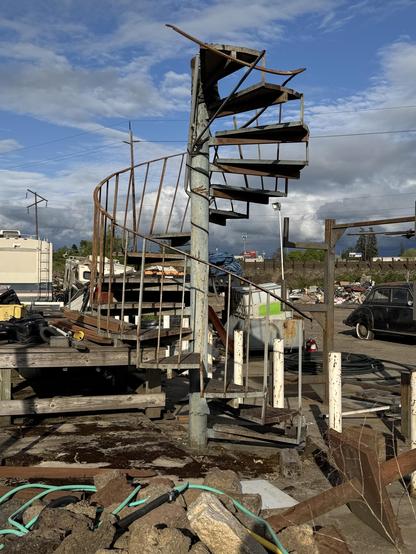 A rusted spiral staircase stands amid debris, with scattered construction materials and an old car in the background. The sky is partly cloudy.