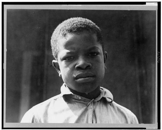 This black and white photograph depicts a young African American male, likely in his late teens or early twenties. He appears to be wearing a light-colored button-up shirt with the top few buttons undone at the collar, giving off an informal look. His hair is short-cropped, closely shaved on the sides but slightly longer on the top. The boy's expression is serious and contemplative as he gazes directly into the camera lens.
The background of the photograph is plain and dark, providing a stark contrast to his lighter skin tone and clothing. There are no discernible objects or features in the backdrop that provide context for the setting where this photo was taken. This image seems reminiscent of works by photographers like Dorothea Lange during her time documenting various social conditions.
Additional information about this photograph can be found through a reference provided: "Delta cooperative farm, Hillhouse, Mississippi," indicating its historical significance as part of agricultural developments or cooperatives in that region during the early 20th century. The name Clarence Weems suggests he was photographed by Dorothea Lange for her project on co-ops and possibly related to evictions from Arkansas due to his father's experiences with beatings and disappearances, highlighting socio-economic challenges faced by African American communities at that time.
The image is part of a collection titled "Dorothea Lange full,"  [...]