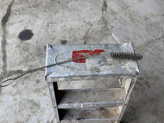 An old, weathered metal step stool rests on a concrete surface. A long, thin, spiral wire brush with a loop handle is placed on top of the stool, alongside a faded red logo or mark. The surrounding area shows signs of wear