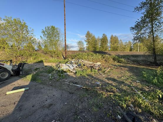 An outdoor area featuring scattered debris and overgrown vegetation. In the foreground, there is a small machinery vehicle, and a telephone pole is visible in the background under a mostly clear sky.