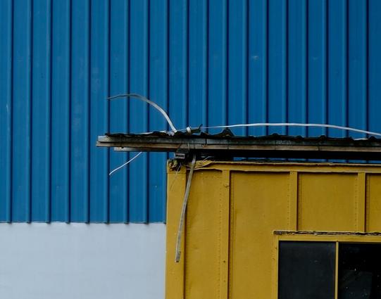 The corner of a yellow corrugated metal workers shed against a wall with blue corrugated cladding and white painted concrete. The roof of the shed is fastened down with metal ties that have not been trimmed and reach out like a claw.