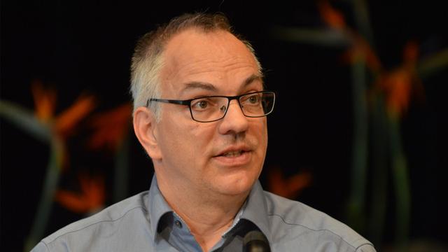 Portrait von Christof Voigt. Ein Mann mittleren Alters mit kurzen, grau melierten Haaren und einer Brille. Er trägt ein hellblaues Hemd und spricht in ein Mikrofon. Der Hintergrund ist unscharf und zeigt orangefarbene Blüten.