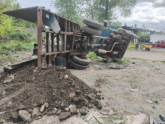 An overturned truck rests on a pile of dirt in a junkyard, surrounded by scattered debris and greenery.