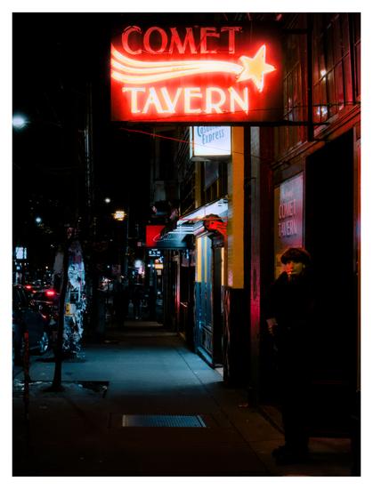 Man smoking a cigarette at night, under a neon sign that says "Comet Tavern".