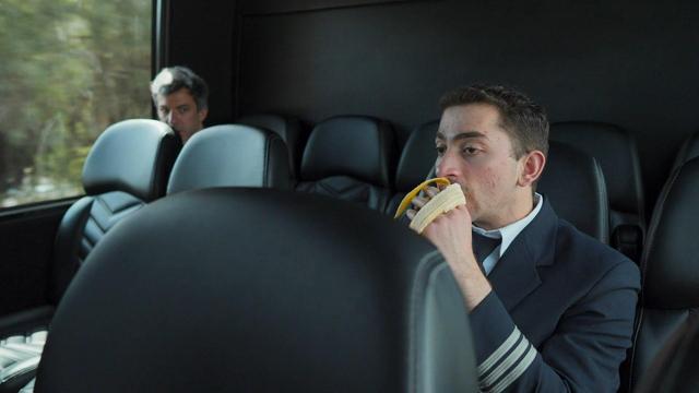 screenshot in a transit but with the camera observing a young man in his pilot uniform eating a banana and nathan sitting a row behind and at the far end, observing the young pilot eating the banana