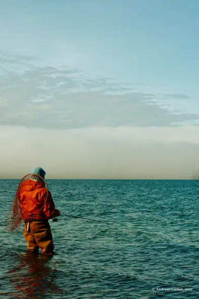 Im blauen Wasser unter einem leicht bewölkten Himmel steht ein Fischer in Wathose, mit dicker Jacke und Mütze im Wasser und hält seine Angel hinaus. Auf dem Rücken trägt der Angler ein großes Netz.