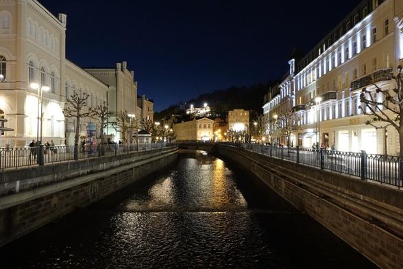 Blick in Karlsbad von einer Brücke auf die Eger am ABend, rechts und links beleuchtete Häuser. im Hintergrund ist ein beleuchtetets Haus auf einem Berg zu sehen.