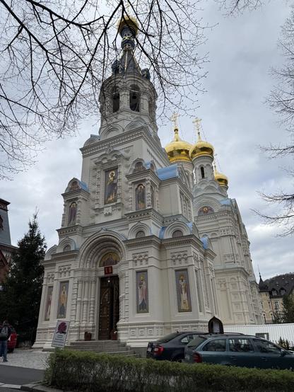 Das Bild zeigt die weiß-blaue orthodoxe Kirche der heiligen Aposten Peter und Paul mit goldenen Kuppeln. Die Kirche ist im traditionellen russischen Stil erbaut und mit zahlreichen Ikonen und Verzierungen geschmückt.  Im Vordergrund stehen einige Autos geparkt und im Hintergrund sind kahlköpfige Bäume und ein weiteres Gebäude zu sehen. Der Himmel ist bewölkt.