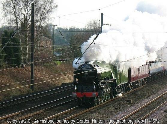 A green steam locomotive drawing, at speed, several carriages along a railway line.  A white plume of steam  from the funnel obscures the cab and part of the. First carriage