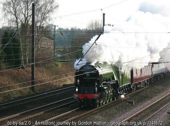 A green steam locomotive drawing, at speed, several carriages along a railway line.  A white plume of steam  from the funnel obscures the cab and part of the. First carriage