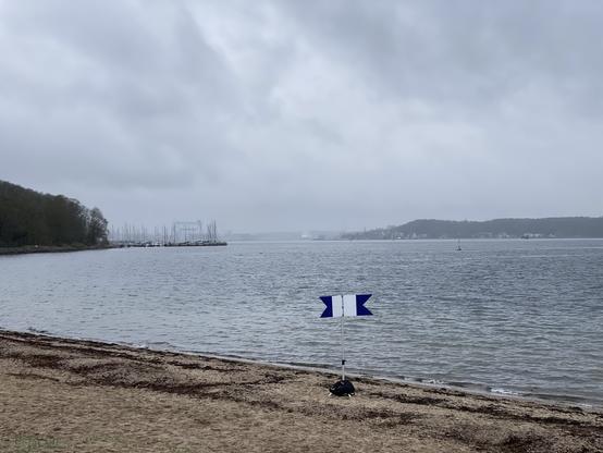 Die Signalflagge "Alpha" (Taucher im Einsatz) steht am Spülsaum des Strandes von Kitzeberg an der Kieler Förde. Im Hintergrund sind die Kieler Skyline mit den Werftkränen durch leichten Nieselregen hindurch noch gerade so zu erahnen. Etwas deutlicher ist die Kiellinie und das Düsternbrooker Gehölz direkt gegenüber zu sehen. In der Nähe des Strandes schwimmt eine gelbe Untiefenwarntonne, etwas weiter draußen fährt eine der Hybridfähren der SFK nach Laboe.