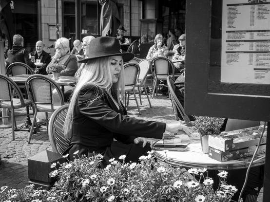 The image depicts a woman sitting at an outdoor café table. She is wearing a black hat and a dark jacket, with long blonde hair cascading down her back. The table in front of her is adorned with a small potted plant, a box, and a smartphone. The setting is a bustling café with other patrons seated at tables, some engaged in conversation. The cobblestone pavement and the arrangement of tables and chairs contribute to the café's inviting atmosphere.