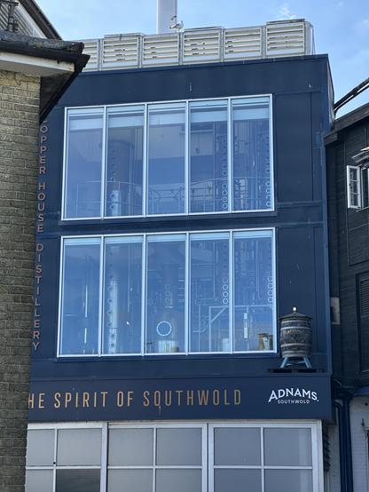 A building of about three floors high. On it is lettering of the spirit of Southwold, and Adnams below a model of a beer keg. Floors two and three are a sequence of tall windows behind which appears to be lots of vertical cylinders with circular ports on them, many clustered together like tentacle suckers.