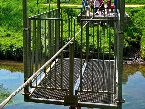 The rectangular gondola of the transporter bridge. A rope runs through its centre. Several people are standing on the other bank. They have just crossed the canal and a child is in the process of sending the gondola back to me. Thank you!