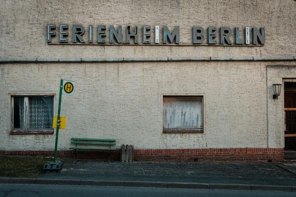 Auf einer patinierten Gebäudefassade prangt ein in die Jahre gekommenes Schild "FERIENHEIM BERLIN". Auf der linken Seite befindet sich ein Fenster mit einem Spitzenvorhang und einer grünen Parkbank unter einem angrenzenden Bushaltestellenschild. Quer über Wand verläuft ein Regenrohr. Neben einer halben Tür hängt eine Latüchte