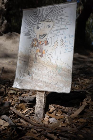 A hand-drawn picture set in a wooded area on a wooden post. The drawing features a whimsical character with large eyes, a wide smile, and exaggerated spiky hair. The figure wears a patterned garment and appears to be waving or holding something in its hand. Handwritten text beside the character includes a date, \"11.11.2023,\" and a name, \"Ronny White.\" Additional text at the bottom reads \"the lost giant of Mandurah.\" The sign is surrounded by leaves and twigs on the ground.