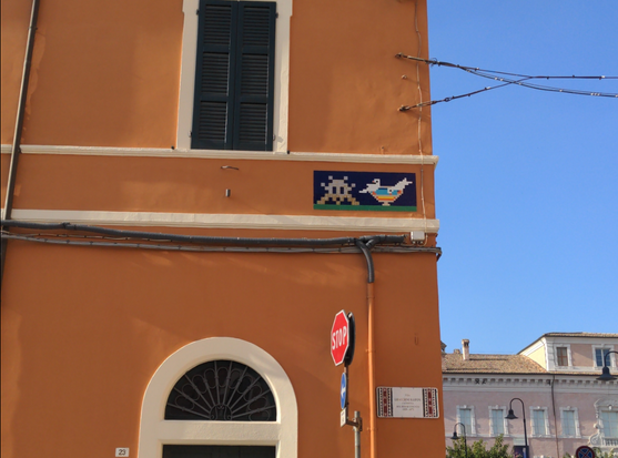 À l'angle d'un bâtiment peint en orange, en mosaïque, un Invader gris sur fond noir, à côté d'un bol multicolore où boivent deux oiseaux, reprenant une mosaïque antique du mausolée de Galla Placidia.